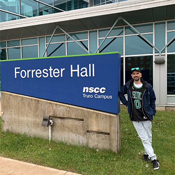 Shaun leans against the NSCC Truro sign in front of a large building.