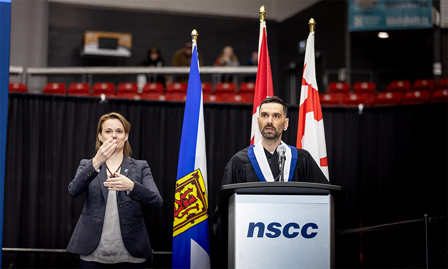 Shaun stands on stage in graduation gown and stole and delivers his speech. An ASL interpreter stands beside him.