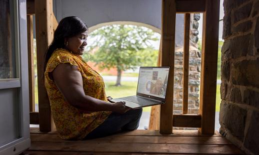 NSCC grad Donna Johnson wears a blouse and jeans and sits on porch stairs while taking part in an online meeting on her open laptop.