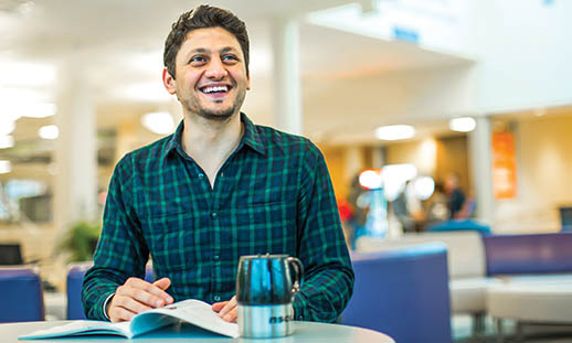 A young man looks off to the top right off the image. He is smiling while sitting at a table. His hand is on a book. He is wearing a button-up plaid shirt. The background is bright and open.