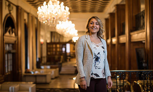 A young woman smiles and looks at the camera. She is wearing a blazer and flowery shirt. She is in a large room with multiple chandeliers, wood paneling and soft seating.