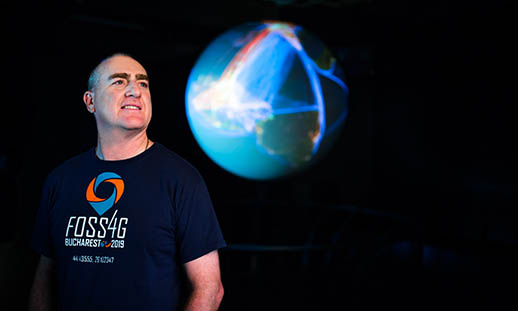 A bald man looks off to the top, right of the image. He is wearing a t-shirt that reads F O S S 4 G Bucharest 2019. Behind him, a well-lit, large globe can be seen.