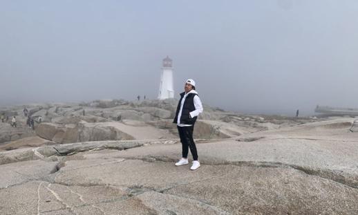 Gabe stands on the rocks with the Peggy's Cove Lighthouse in the middle distance behind him.