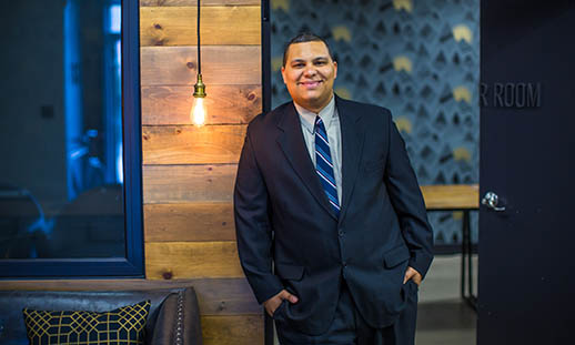 A young man leans against a door casing while smiling at the camera. He is wearing a blue suit and tie. There is a wood clad wall and exposed lightbulb to the left. Behind the man is a room with modern wallpaper.