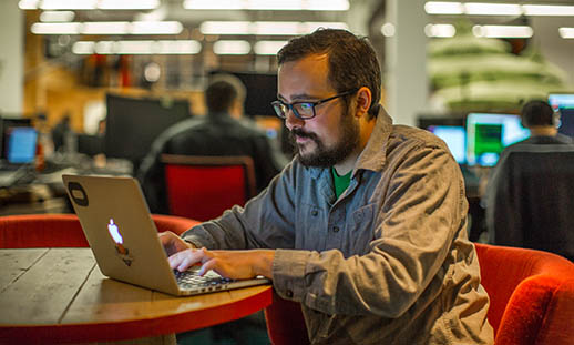 A bearded man wearing glasses sits in a red chair at a round desk and works on an Apple laptop. Behind him, other people doing similar work can be seen.