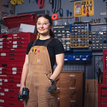 A female carpentry student smiles at the camera. She is wearing safety glasses and Carhart overalls while standing in front of multiple tool boxes in a workshop.