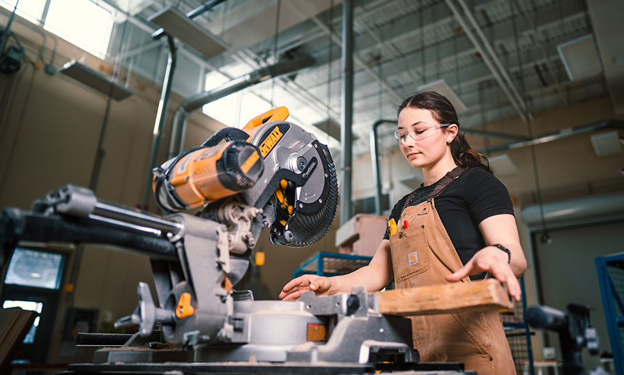A female carpentry student prepares a piece of wood before cutting it. She is wearing clear safety glasses and brown Carhart overalls with some tools in her front pocket.