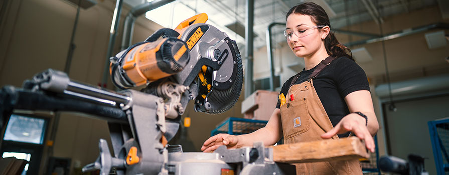 A female carpentry student prepares a piece of wood before cutting it. She is wearing clear safety glasses and brown Carhart overalls with some tools in her front pocket.