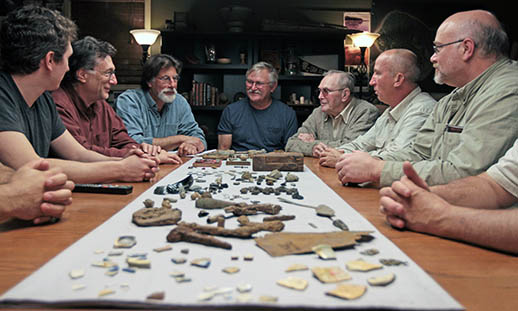 Alex Lagina, Marty Lagina, Rick Lagina, Dave Blankenship, the late Dan Blankenship, Gary Drayton and Doug Crowell all sit around the head of a long table on the set of the History Channel show The Curse of Oak Island. On the table, dozens of pieces of wood, potter, bone and metal are laid out. Most men are looking to Rick Lagina, who appears to be talking. 