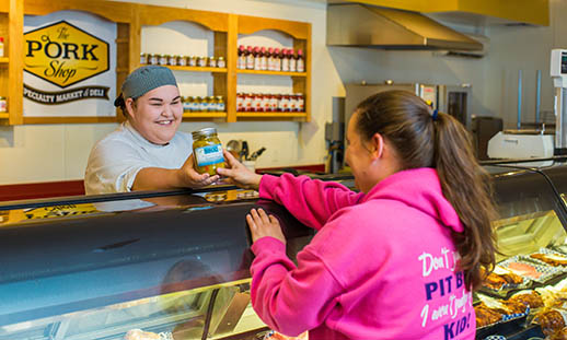 A young woman wearing a chef jacket and pill-box cap, stands behind a tall, deli counter and passes a jar to a woman in a pink sweater whose face is not seen.