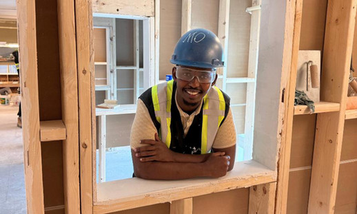 Smiling man wearing a hard hat and safety glasses, standing in a window frame inside a woodworking shop