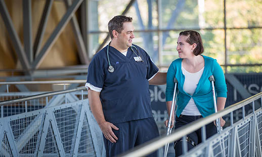 A man in blue scrubs with a stethoscope around his neck, walks down a railed walkway with a woman who is using crutches. 
