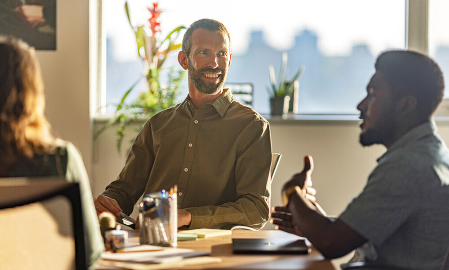 Joe sits at a board room table in front of a window and smiles at the person speaking.