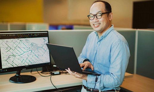 Justin Chang, smiling while holding a laptop and standing next to a screen with a map on it