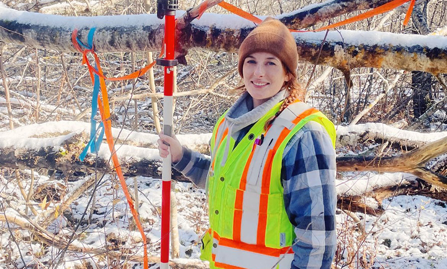 A woman wearing a safety vest stands in a snowy forest holding surveying equipment