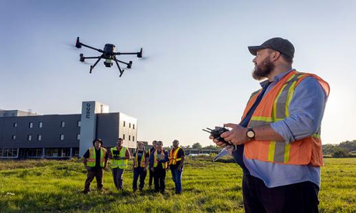 Student wearing safety vest uses remote to fly drone while students watch nearby