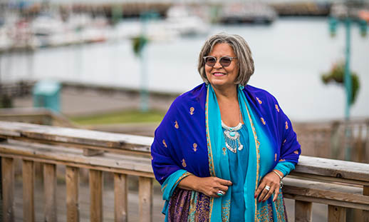 An woman wears a bright, multi-blue-coloured jacket and looks off the left of the image. She is on a wooden walkway and leans up against the railing. Water and boats can be seen in the background.