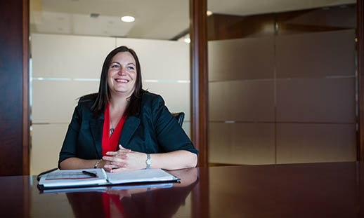 A woman smiles and looks off to the right of the image. She is sitting at a large, brown table and wears a dark blazer and red, v-neck shirt. There is an open notebook on the table in front of her. 