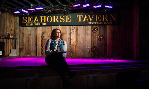 A woman sits on a stage wearing a jean vest and white shirt. She is looking off to the right. The stage wall is clad in rough lumber and a sign reads the seahorse tavern. A pink-purple light is shining on the floor. 