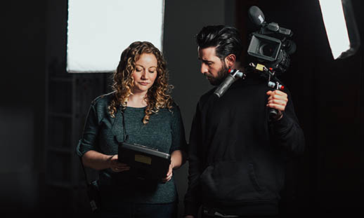 A woman with blonde, curly hair points to an iPad while showing the screen to a man holding a large video camera on his shoulder. The woman is wearing a green sweater and the man is dressed in all black. There are large, square lights seen in the background. 