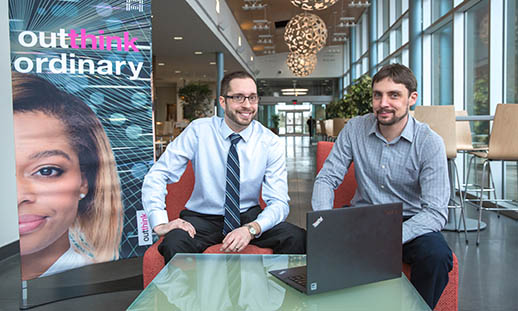 Two young men sit on chairs and look at the camera. They are wearing buttoned up shirts. On a low, glass table in front of them is a black laptop. There is a pull-up banner to the left that reads: IBM outthink ordinary.