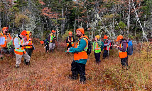 A group of Natural Resources Environmental Technology students stand outdoors in a forested area wearing safety vests and holding clipboards. Mitchell stands in the centre of the photo.