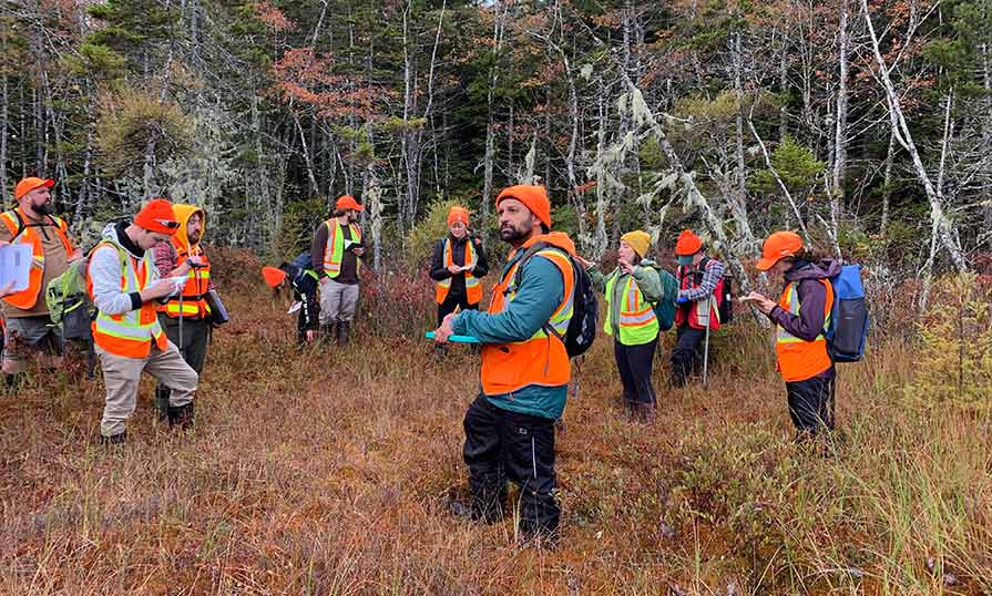 A group of Natural Resources Environmental Technology students stand outdoors in a forested area wearing safety vests and holding clipboards. Mitchell stands in the centre of the photo.