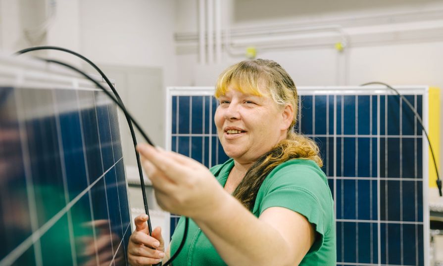 A woman standing between two solar panels holding two black wires.  