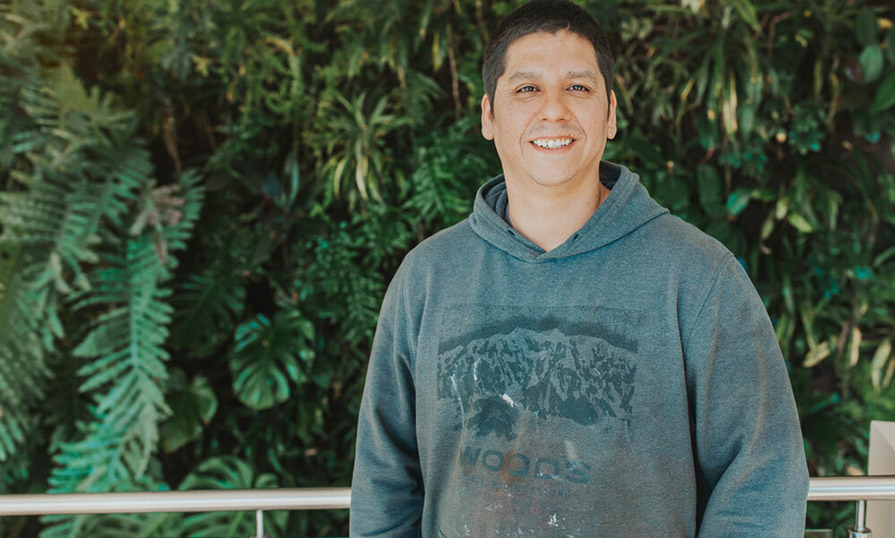 A student wearing a grey hoodie stands in front of a wall covered in foliage.