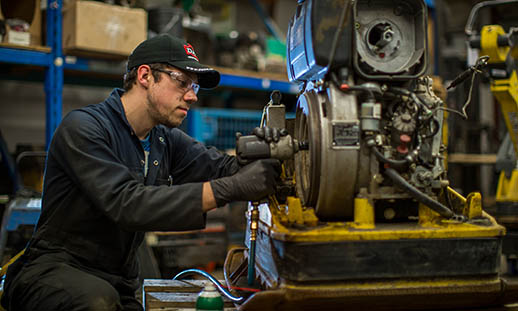 A man in blue coveralls, a ball cap, safety glasses and black gloves works on a mechanical item. He is in an industrial shop environment. 