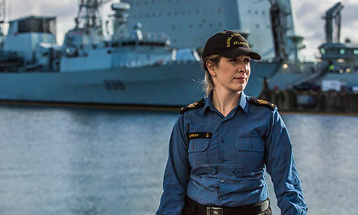 A woman, wearing a Canadian Navy uniform and black cap, looks off to the right. In the background is water and two navy ships.