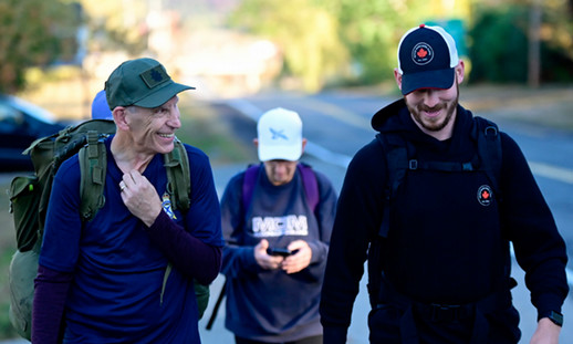 Image shows two people walking. One is wearing a black hoodie with a Canadian flag and the other is wearing a military uniform carrying a ruck sack.