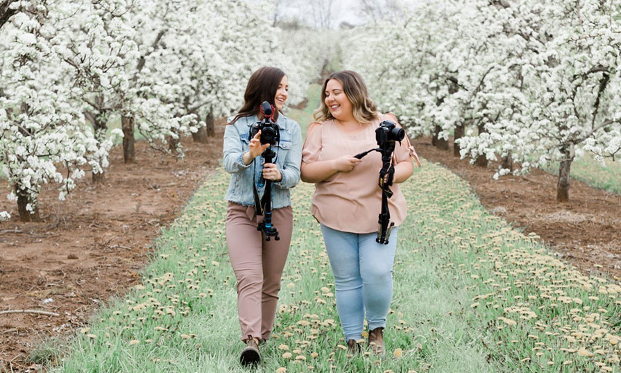 Two women walk in a flowery field carrying camera equipment.