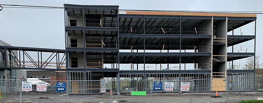 The metal framing of a new building is under construction. The weather is cloudy and the construction site is fenced in.