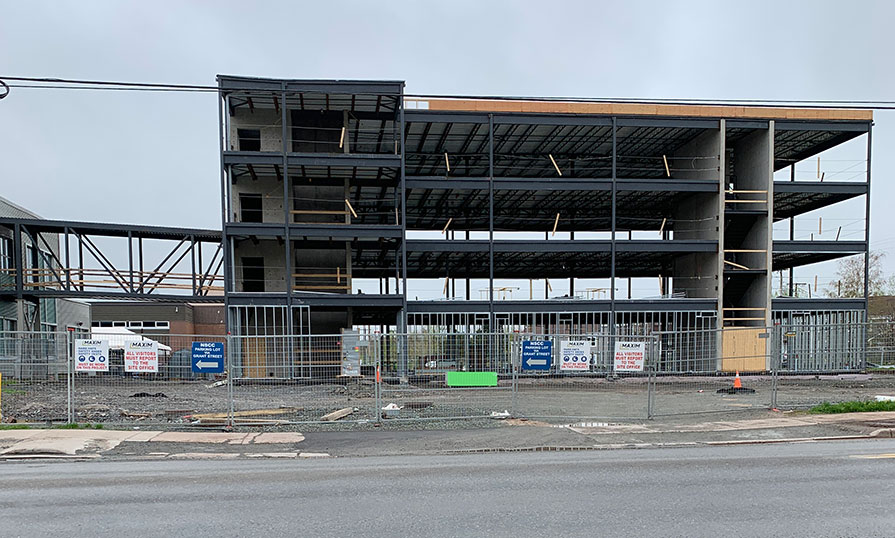 The metal framing of a new building is under construction. The weather is cloudy and the construction site is fenced in.