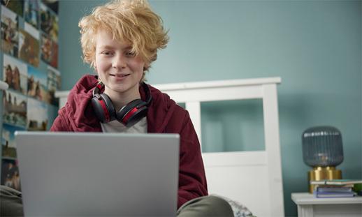 Teenager with short, curly blonde hair sits on a bed watching a session on a laptop.