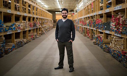 A young man with a beard and glasses, wearing a v-kneck sweater and white t-shirt, stands in the center of an isle in a warehouse. The shelves hold many kinds of wooden planks.