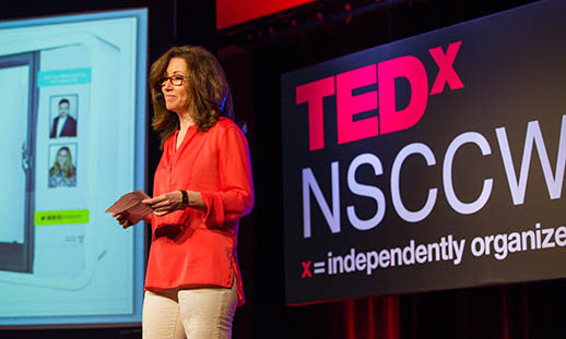 A woman in an orange shirt and glasses stands in front of a large sign that reads TEDx NSCC Wa…(the rest is unseen). She is holding cue cards in her hands and appears to be giving a presentation.