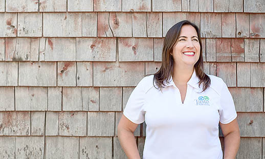 A woman in a white t-shirt smiles while leaning against a wooden shingled wall and looking to the right. On her shirt, the word taste can be read. 