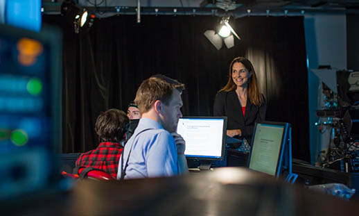 Three individuals are shown in a television studio. It is dark. Two are seated at computers with their backs to the camera. A third person, standing, is looking at the two seated people and smiling. 