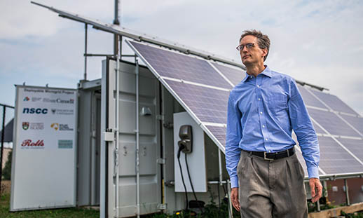 A man in a blue, buttoned up shirt and khaki pants walks in front of a large section of solar panels that are attached to a white shipping container. He is outside.