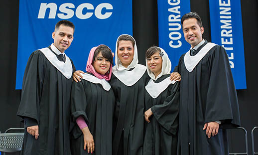 Five people stand shoulder to shoulder in black graduation gowns and white stoles. Three women in the centre wear hijabs and the men at each end wear collared shirts and ties. In the background, fabric signs read N S C C, courage and determined.