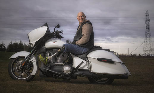 A man smiles while sitting astride a white motorcycle. He is wearing jeans, a brown sweater and a vest. His leg is a prosthetic, and he is wearing a bright green sneaker. There is a large power line in the background.