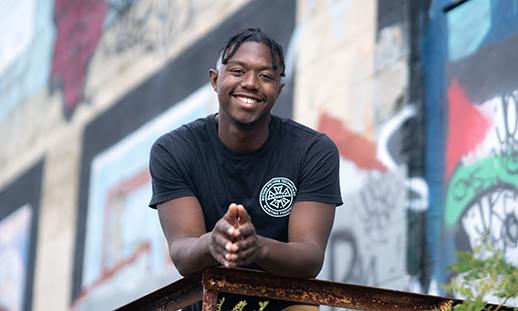 Keandre Johnson, an NSCC graduate, stands in front of a brick building covered in graffiti and leans forward on a railing while smiling.
