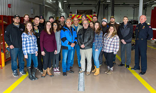 A group of 21 individuals stands close together and smiles for a photo in a firehouse. While most appear to be students, the two on the far right are not. Both are dressed in business attire. One is dressed in a uniform.