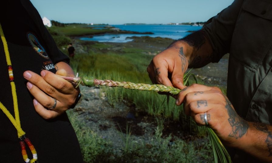 Two hands, one adorned with tattoos, collaboratively braiding long strands of grass on a marshy coastline. 