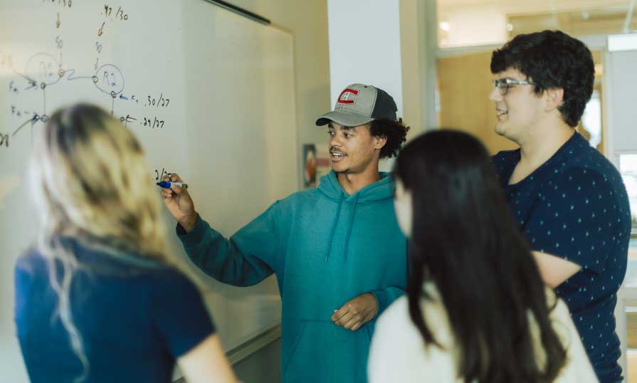 Students gathered at the front of the class standing beside a whiteboard while one student is explaining the graph that is shown on the board