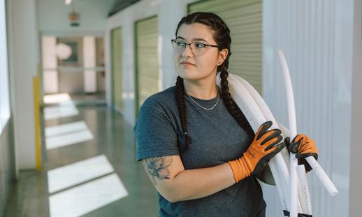 Taylor O'Hearn wears glasses, a t-shirt and safety gloves and carries a role of plastic pipe over her left shoulder while gazing to her right.