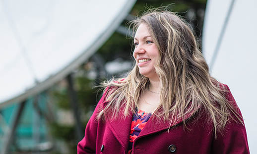 A blonde woman smiles and looks to the left of the image. She is wearing a wine-coloured pea coat. In the background, two, large satellite dishes can be seen.