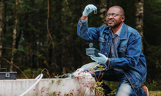 A man crouches by a well. He is wearing a jean jacket and blue latex gloves. He is holding a test tube and looking at it. In the background there is a thick crop of trees.
